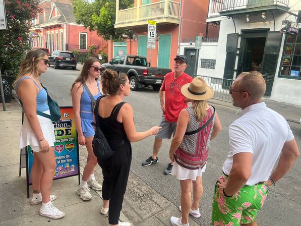 Six people chatting on a sunny historic French Quarter street with pastel Creole-style houses, iron balconies, parked trucks, and a colorful sidewalk sign