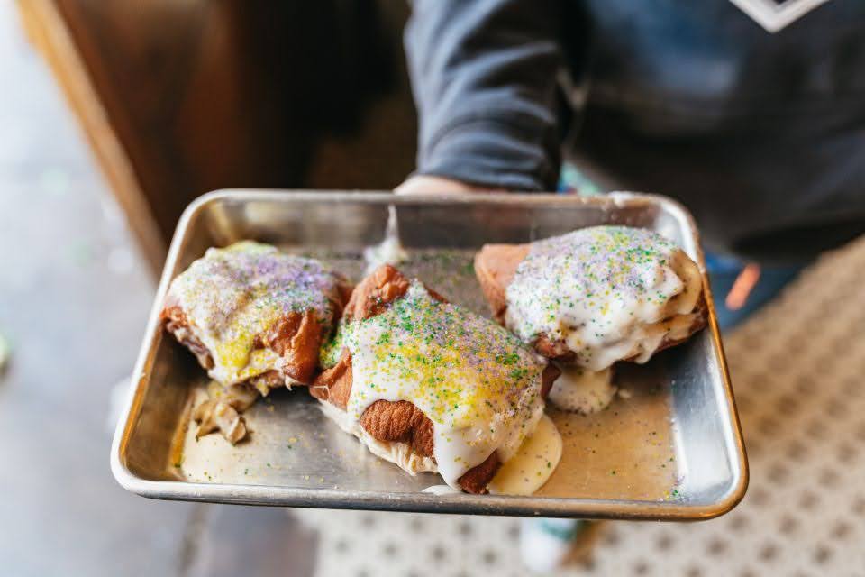 Three warm cinnamon rolls with white glaze and purple, green and gold Mardi Gras sprinkles on a metal tray held by a server.