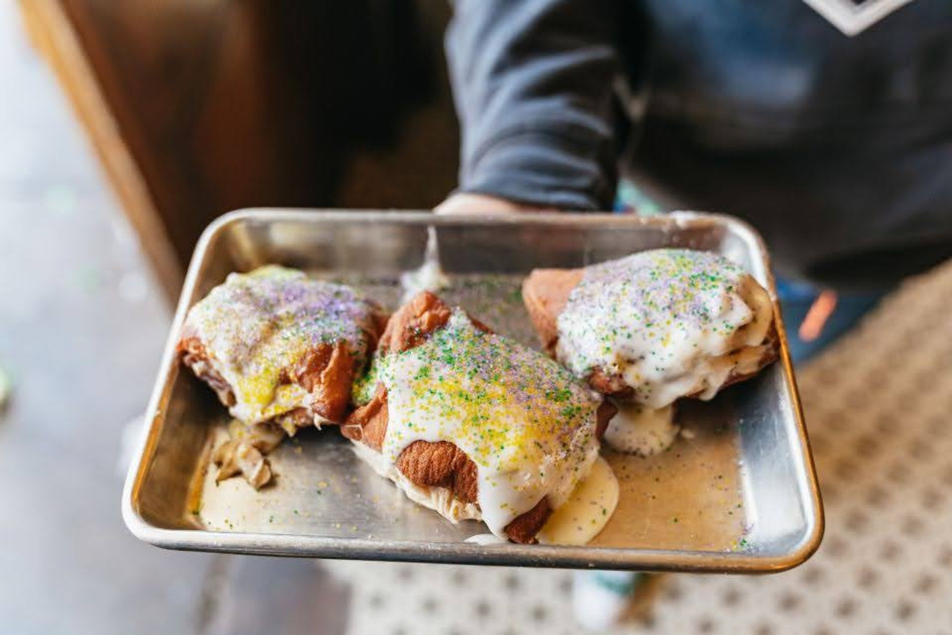 Three warm cinnamon rolls with white glaze and purple, green and gold Mardi Gras sprinkles on a metal tray held by a server.