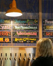 Craft brewery taproom interior with stainless beer taps and a colorful chalkboard menu under an orange pendant light listing playful beers like Watermelon, That Mother Shucker, and Retro Cartoons.