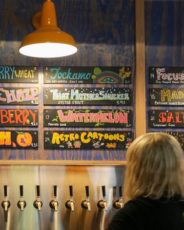 Craft brewery taproom interior with stainless beer taps and a colorful chalkboard menu under an orange pendant light listing playful beers like Watermelon, That Mother Shucker, and Retro Cartoons.