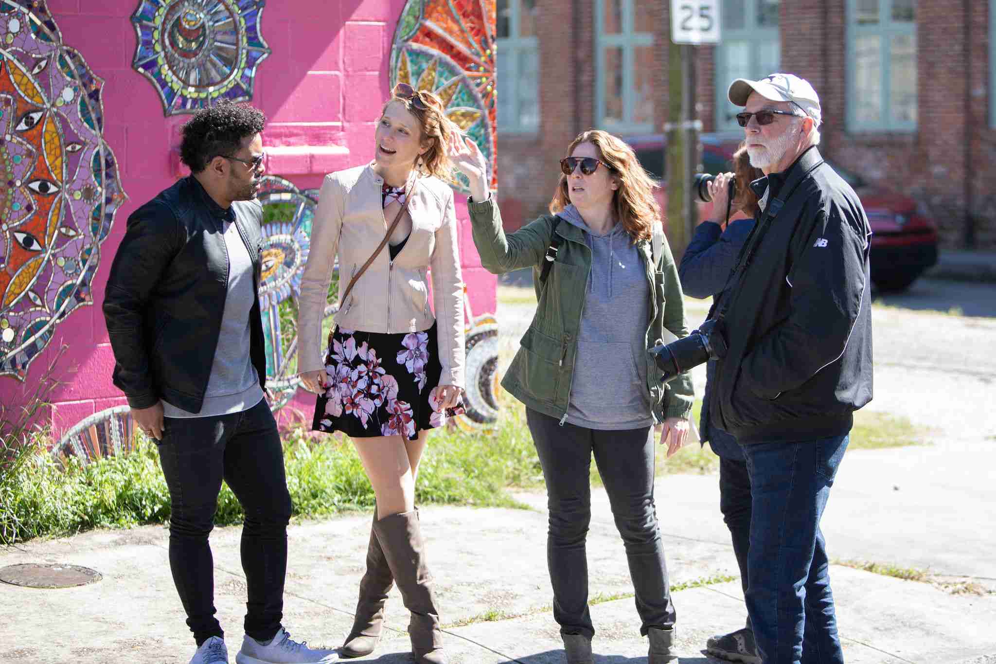 Four adults on a sunny urban sidewalk beside a colorful mosaic street-art mural on a pink wall; two women and two men chatting—one woman pointing, one man holding a camera.