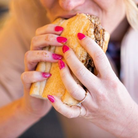 Close-up of hands with bright pink glitter nails and rings biting into a large shredded beef sandwich on a crusty roll — messy savory bite.