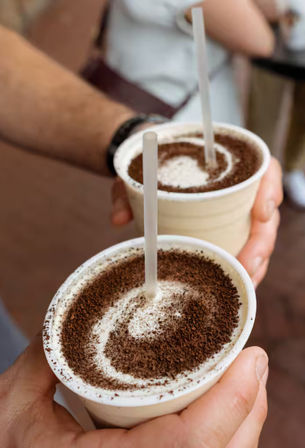 Two creamy chocolate milkshakes in white to-go cups topped with cocoa swirls and clear straws, held in hands