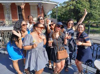 Group of women laughing and posing with cocktails on a sunny rooftop balcony featuring decorative iron railings and a historic brick facade, outdoor daytime celebration near a park.