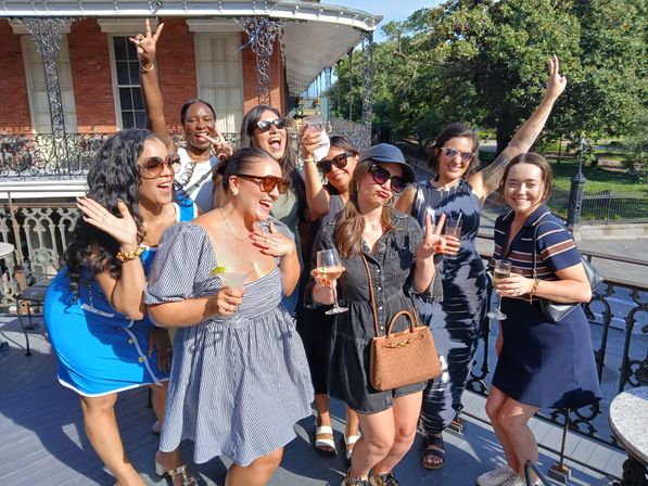 Group of women laughing and posing with cocktails on a sunny rooftop balcony featuring decorative iron railings and a historic brick facade, outdoor daytime celebration near a park.
