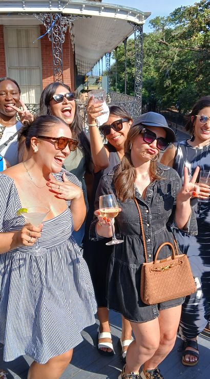 Group of women laughing and posing with cocktails on a sunny rooftop balcony featuring decorative iron railings and a historic brick facade, outdoor daytime celebration near a park.