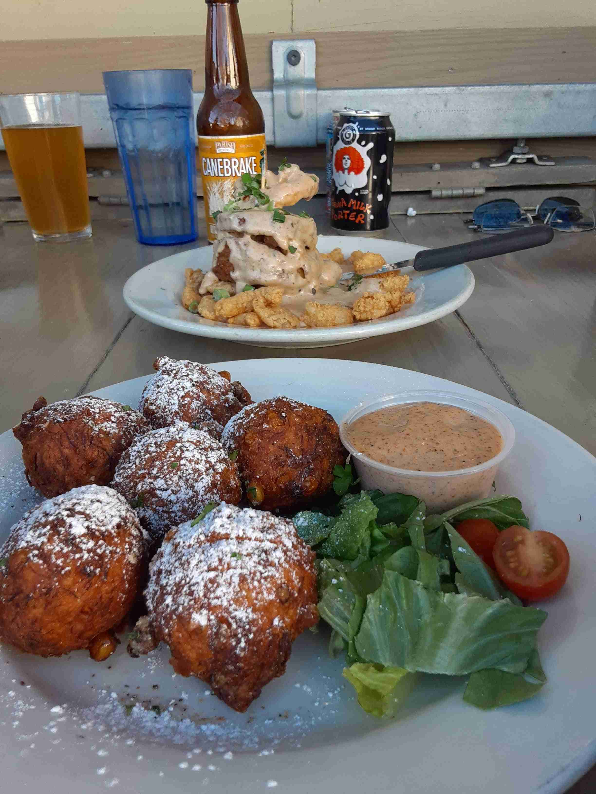 Close-up of a plate of crispy fried fritters dusted with powdered sugar, served with mixed greens, cherry tomatoes and a dipping sauce on a casual outdoor patio table; in the background another plate of fried seafood smothered in creamy gravy with a beer bottle and can nearby.