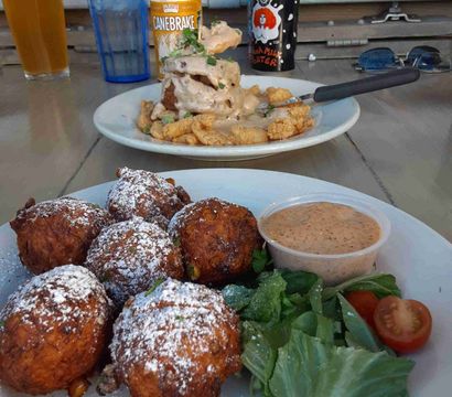 Close-up of a plate of crispy fried fritters dusted with powdered sugar, served with mixed greens, cherry tomatoes and a dipping sauce on a casual outdoor patio table; in the background another plate of fried seafood smothered in creamy gravy with a beer bottle and can nearby.