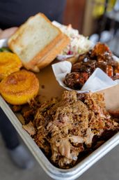 Mouthwatering close-up of a Southern-style barbecue tray with shredded pulled pork, saucy ribs in a paper cup, toasted white bread, golden cornbread rounds, and creamy coleslaw