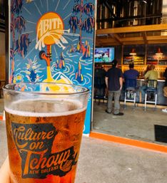 Cold amber craft beer in a pint glass stamped "New Orleans" in the foreground, colorful mural behind and patrons at a casual taproom bar in the background.