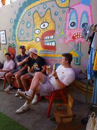 Four friends relaxing on an outdoor urban patio, eating popsicles and sipping beer in front of a bright, whimsical street-art mural of cartoon faces.