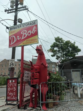 Retro red-and-yellow storage and art sign on a pole beside a tall red metal creature sculpture with attached mailboxes at a fenced urban art gallery entrance, potted plants, speed-limit sign and overhead power lines under a cloudy sky.