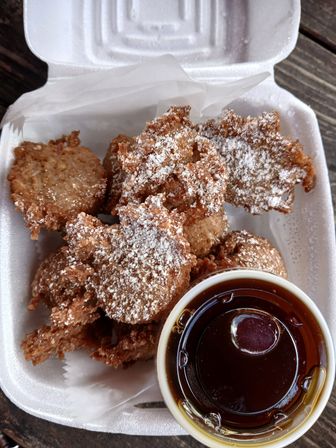 Crispy golden-brown fritters dusted with powdered sugar in a white takeout box, served with a small cup of dark maple syrup on a wooden table.