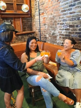 Three friends laughing and toasting bright orange cocktails in a cozy brick-walled bar booth