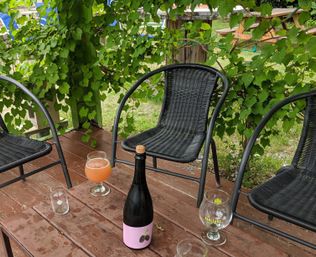 Cozy backyard deck with black wicker chairs under a grapevine pergola, wooden picnic table holding a bottle with a pink berry label, a hazy peach-colored beer in a tulip glass and empty tasting glasses.