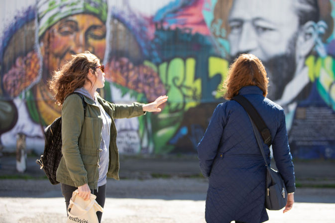 Two women in jackets stop on a sunlit city street to admire a colorful urban mural of large painted faces, one gesturing toward the street art.