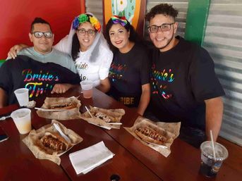 Four friends smiling at a wooden table in a casual eatery, celebrating a bachelorette — bride-to-be in a veil and flower crown with others in rainbow “Bride’s Tribe” shirts, sharing loaded hot dogs and drinks.