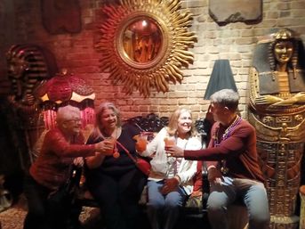 Four people clinking cocktails on a vintage sofa in an Egyptian-themed bar lounge with exposed brick, a sunburst mirror, ornate lamp and sarcophagus decor, wearing festive beads.