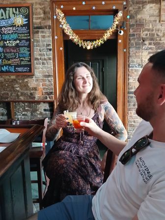 Two people clinking cocktails at a cozy brick-walled bar — smiling tattooed woman in a floral dress and a man in a white shirt, festive garland and chalkboard menu in the background.