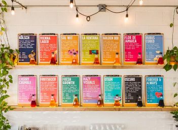 Bright brewery taproom scene with two shelves of colorful craft beer menu boards on clipboards, tasting pours in small bottles, string lights and hanging plants