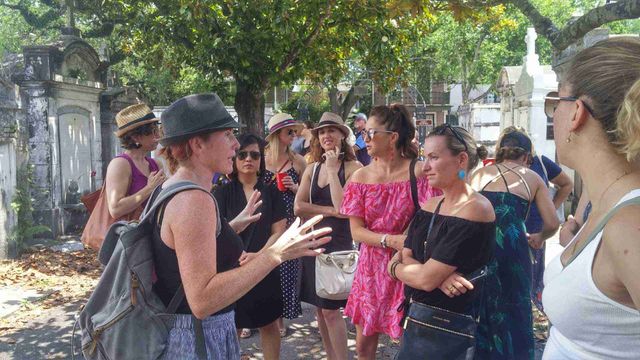 Tour guide animatedly addressing a group of women on a sunny walking tour amid mossy above‑ground tombs and oak trees in a historic cemetery