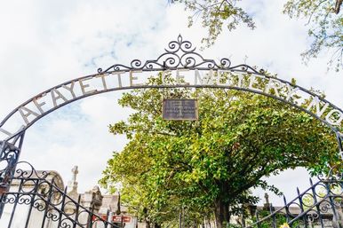 Ornate wrought-iron arch reading "Lafayette Cemetery" over an iron gate, with a large magnolia tree and historic above-ground tombs under a cloudy sky