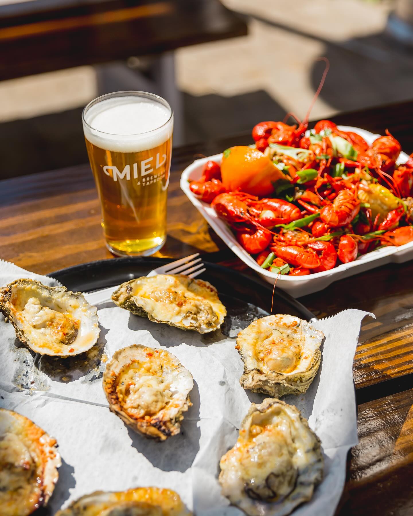 Outdoor wooden table with baked oysters on the half shell, a tray of bright red boiled crawfish with a lemon, and a cold pint of craft beer