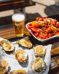 Outdoor wooden table with baked oysters on the half shell, a tray of bright red boiled crawfish with a lemon, and a cold pint of craft beer