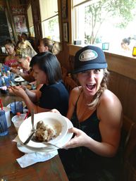 Excited woman in a trucker hat holding a bowl of saucy dessert at a long wooden table during a lively brunch with friends in a sunlit casual cafe.