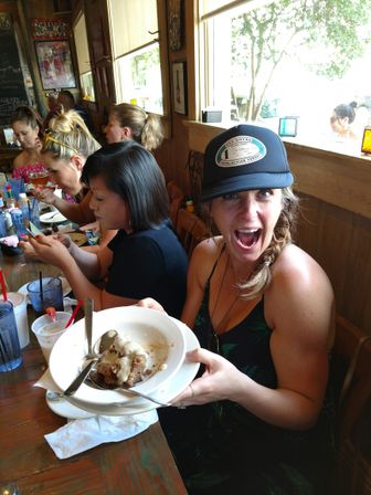 Excited woman in a trucker hat holding a bowl of saucy dessert at a long wooden table during a lively brunch with friends in a sunlit casual cafe.