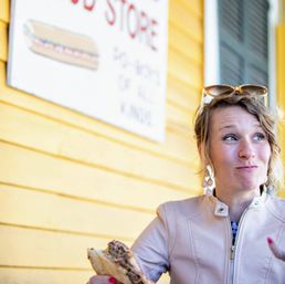 Woman with sunglasses on her head enjoying a shredded beef sandwich outside a bright yellow storefront at a casual deli