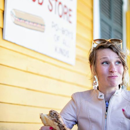 Woman with sunglasses on her head enjoying a shredded beef sandwich outside a bright yellow storefront at a casual deli