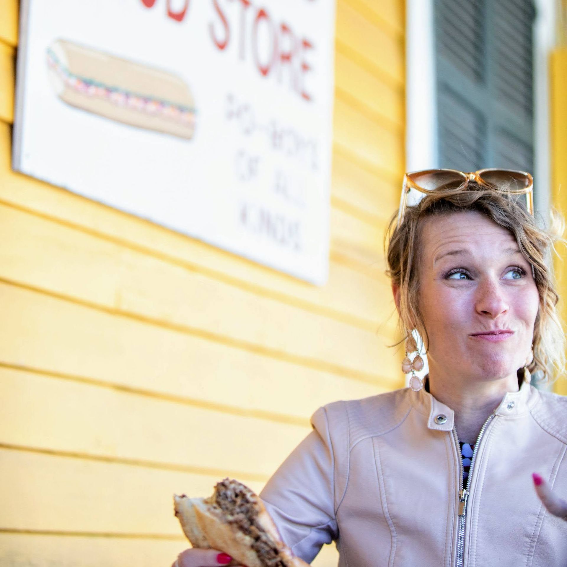 Woman with sunglasses on her head enjoying a shredded beef sandwich outside a bright yellow storefront at a casual deli