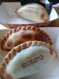 Close-up of three baked Cajun-style hand pies (empanadas) in paper trays; front pie stamped "GUMBO" with golden, crimped crust and browned top.