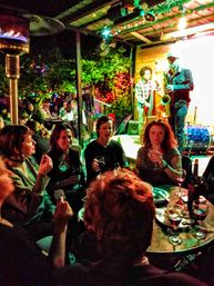 Outdoor nighttime patio with live jazz musicians (saxophone and upright bass) on a small lit stage, friends gathered at a table enjoying wine and appetizers under colorful string lights and greenery.