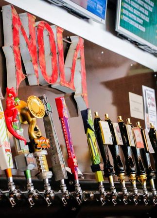 Close-up of colorful craft beer tap handles lined up on a bar draft system with brass faucets and a large red vintage letter sign in the background, ready to pour.