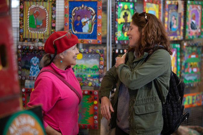 Two women smiling and chatting at an indoor art market booth filled with vibrant folk-art paintings; one in a red beret and pink jacket, the other in a green jacket with a backpack.