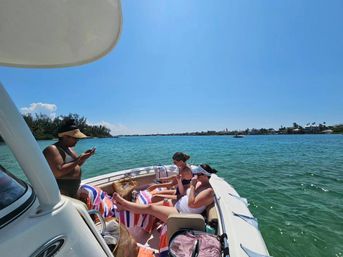 Three people relaxing on a small motorboat in turquoise coastal waters on a sunny day, one using a phone and two lounging on striped towels with a palm-lined shoreline and houses in the distance.