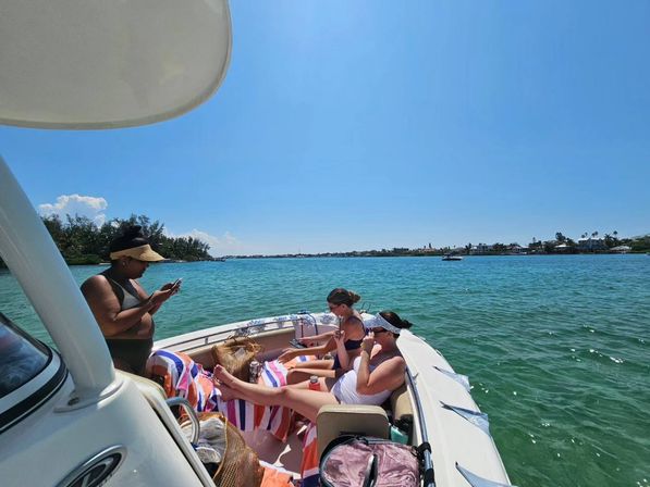 Three people relaxing on a small motorboat in turquoise coastal waters on a sunny day, one using a phone and two lounging on striped towels with a palm-lined shoreline and houses in the distance.