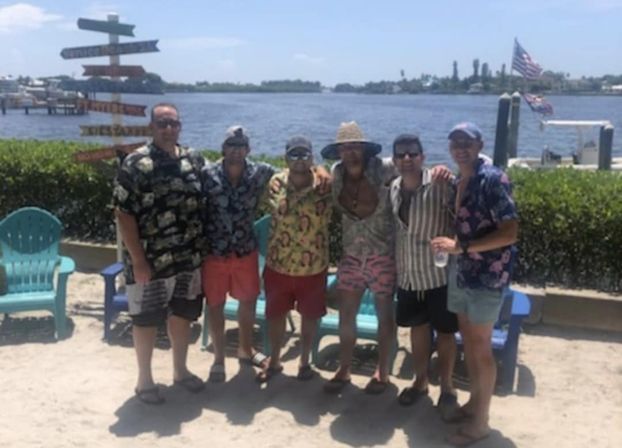 Six men in colorful Hawaiian shirts and shorts posing on a sunny sandy waterfront with turquoise Adirondack chairs, a wooden signpost, dock and American flag in the background.