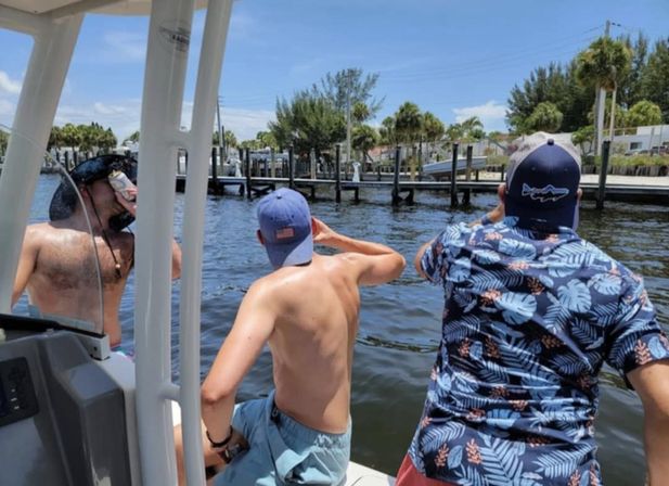 Three people on a small boat in a sunny coastal canal, two shirtless and one in a blue tropical shirt wearing caps, raising drinks and looking toward palm‑lined docks and a marina.