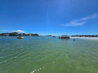 Sunny coastal inlet with shallow turquoise water and a sandy sandbar, small boats anchored near shore and people wading under a clear blue sky