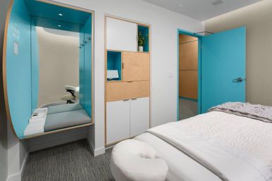 Bright modern spa treatment room with turquoise accents: massage table with face cradle, cozy built-in turquoise seating nook, light wood storage cabinets and open turquoise door in a minimalist wellness interior.
