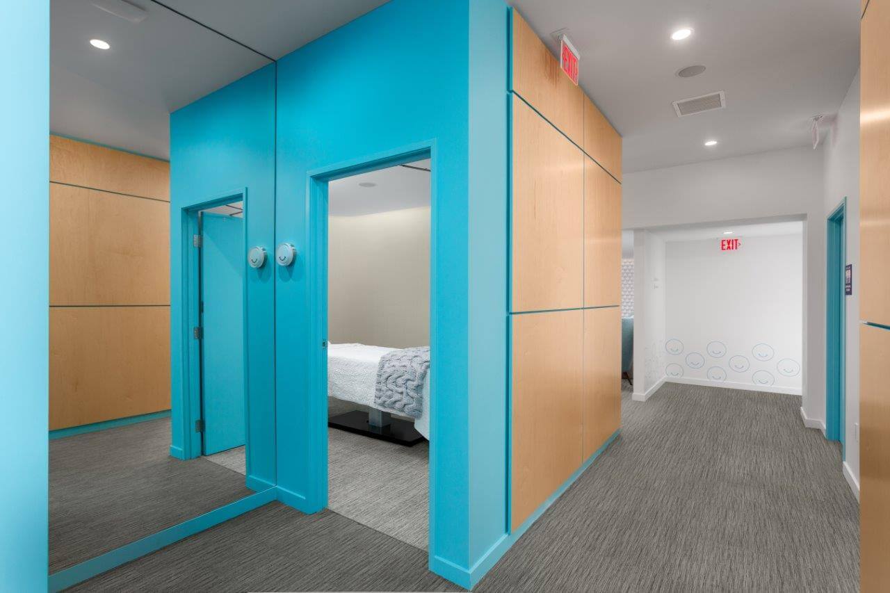 Contemporary interior corridor with bright turquoise doorways, light wood wall panels, gray carpet and a small bedroom visible with a white bed and textured throw, clean modern design with EXIT sign