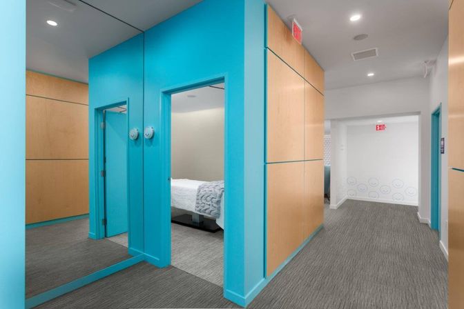 Contemporary interior corridor with bright turquoise doorways, light wood wall panels, gray carpet and a small bedroom visible with a white bed and textured throw, clean modern design with EXIT sign