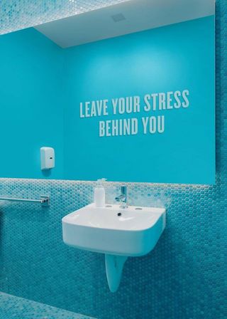 Bright turquoise spa-style restroom with white wall-mounted sink, penny-tile backsplash, soap dispenser and motivational wall text reading "LEAVE YOUR STRESS BEHIND YOU".