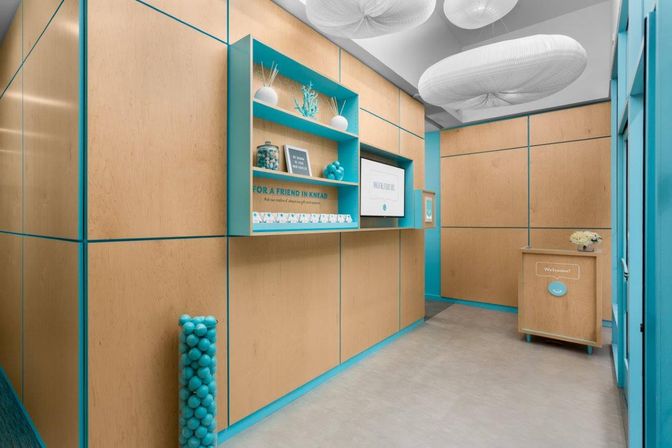 Bright modern reception area with light birch wood-paneled walls and playful turquoise trim, built-in shelves with decorative jars and signage, small welcome desk, tall column of turquoise balls, and oversized soft white pendant ceiling lights.