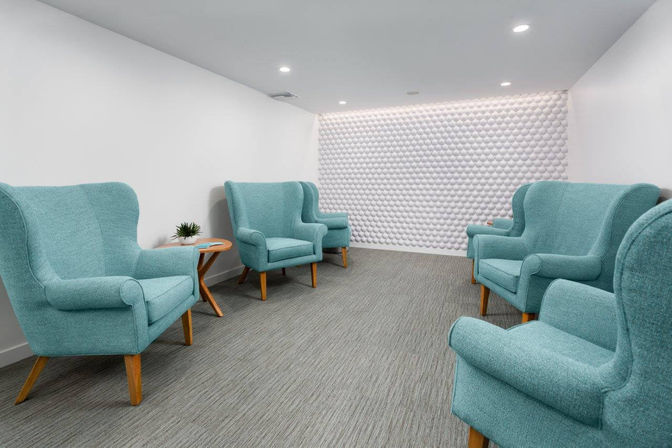 Bright modern waiting room with six teal wingback chairs, wooden side tables, carpeted floor and a white pebble-textured accent wall