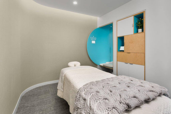 Contemporary spa treatment room with a plush massage table in white linens and a chunky gray knit throw, curved beige walls, a turquoise circular alcove with bench, and built-in wood-and-white storage with a small potted plant.